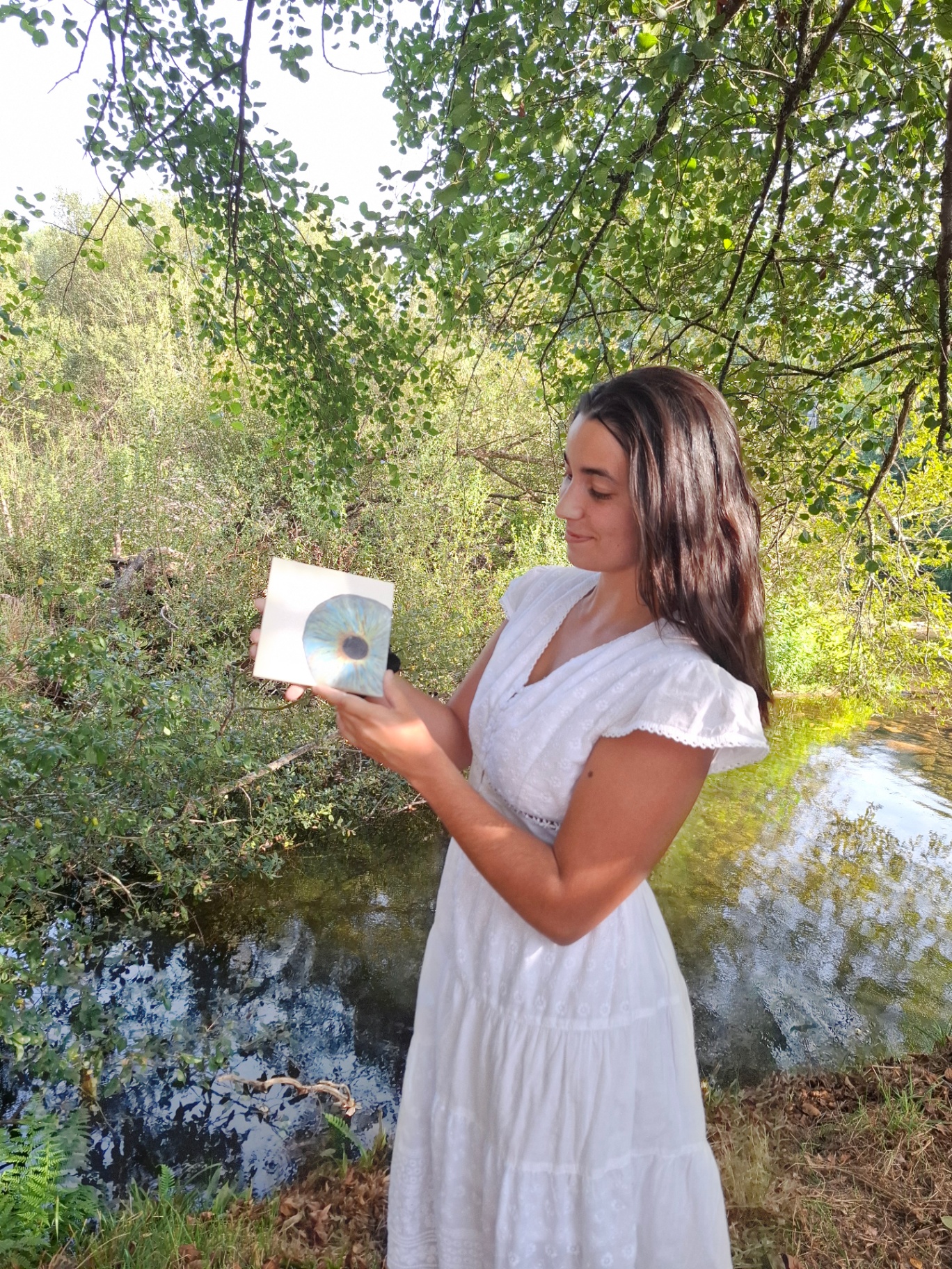 Mulher de branco junto ao rio a segurar um azulejo pintado com um olho